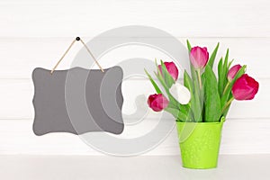 Hanging small black vintage chalkboard on white painted wooden wall near tulip flower bouquet. Spring mockup background