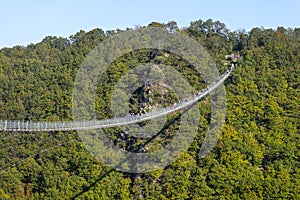 A hanging wooden bridge with steel ropes seen from the side against a blue sky, visible tourists on the bridge.