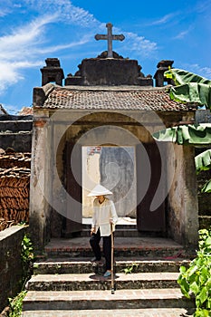 Hanoi, Vietnam - July 17, 2016: Aged church gate with Holy cross on top, Vietnamese old woman wear conical hat and stick walking i