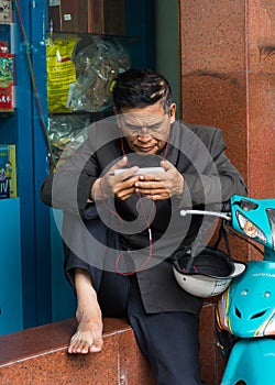 Hanoi, Vietnam - Nov 16, 2014: Senior man reads news and listens to music by earphone from a smartphone on sidewalk on Hang Khay s