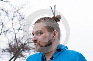 Hansome bearded man with hair bun with sticks and make up looking away in front of tree