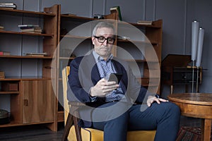 Hansome elderly businessman in blue suit and glasses working and looking on his mobile phone on office wall with boockshelf