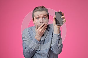 Hansome guy in blue shirt presenting smart phone covering his mouth in shock