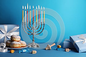 Hanukkah celebration. Menorah with burning candles, dreidels, donuts and gift boxes on light blue table, space for text