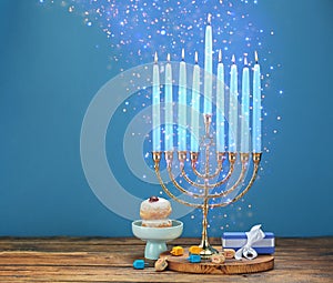 Hanukkah celebration. Menorah with burning candles, dreidels, gift box and donuts on wooden table against light blue background,