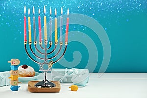 Hanukkah celebration. Menorah with burning candles, dreidels, gift boxes and donut on white wooden table against light blue