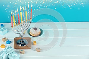 Hanukkah celebration. Menorah with burning candles, dreidels, gift boxes and donut on white wooden table, space for text