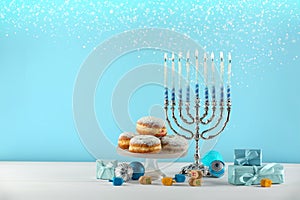 Hanukkah celebration. Menorah with burning candles, dreidels, gift boxes and donuts on white wooden table against light blue