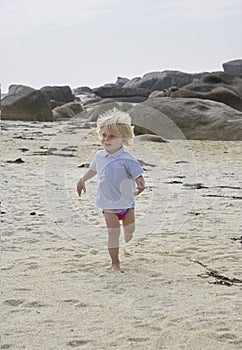 Happily smiling cute little boy running in the sand at a beach, summer holiday feeling, rocks in the background