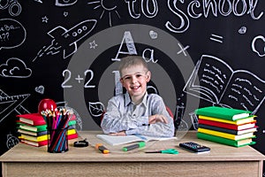 Happily smiling excellent schoolboy sitting on the desk with books, school supplies, with both arms leaned one to