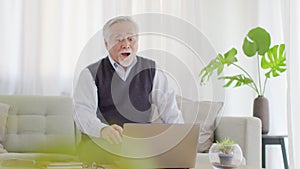 Happiness elderly asian man sitting on sofa and using computer laptop working online with business team and celebrating smile