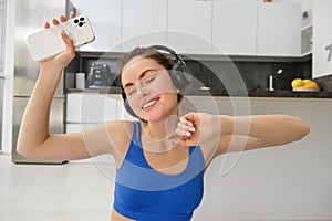 Happiness, sports and wellbeing. Young woman dancing in headphones, holding smartphone, doing workout at home, fitness