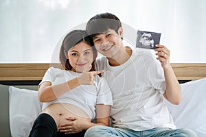 Happiness young Asian couple wearing a casual dress smiling and sitting on a bed together. A happy husband puts his arm around his