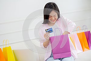 Happiness young asian woman sitting on sofa open paper bag while online shopping with credit card at home.