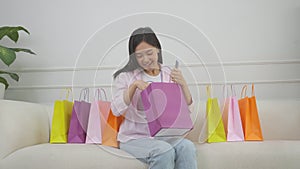 Happiness young asian woman sitting on sofa open paper bag while online shopping with credit card at home.