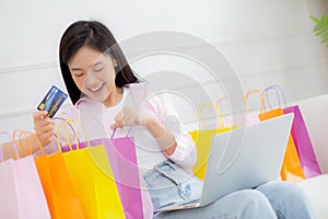 Happiness young asian woman sitting on sofa open paper bag while online shopping with credit card at home.