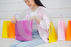 Happiness young asian woman sitting on sofa open paper bag while online shopping with credit card at home.