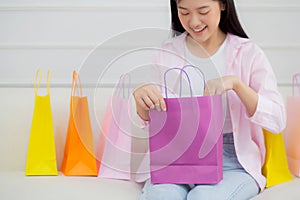 Happiness young asian woman sitting on sofa open paper bag while online shopping with credit card at home.