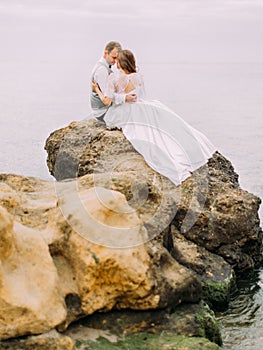 Happpy newlyweds are sitting on the cliff among the sea. The back view.