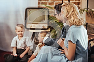 happy 1950s style parents sitting on sofa and lookign at cute little children