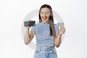 Happy 20s woman shows horizontal mobile phone screen with credit card, laughing and smiling, girl looks excited at