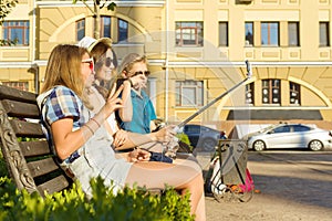 Happy 4 teenage friends or high school students are having fun, talking, reading phone, making selfie photo in city on bench.