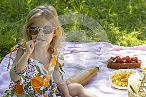 A happy 5-year-old girl is sitting on a mat and enjoying cookies at a picnic in the park. Enjoying outdoor recreation
