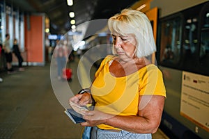 Happy 50s senior woman biometric passport and train ticket waiting train on station platform. Railroad transport concept