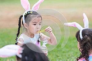 Happy 6 year old Asian little girl with bunny ears blowing soap bubbles in park, having fun with friends, sunny day. Cute children