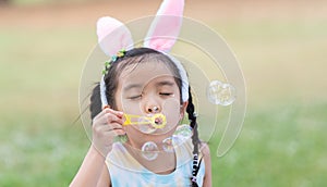 Happy 6 year old Asian little girl with bunny ears blowing soap bubbles in park, having fun, sunny day. Portrait of cute child