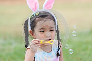 Happy 6 year old Asian little girl with bunny ears blowing soap bubbles in park, having fun, sunny day. Portrait of cute child