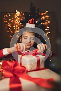 Happy 6 years old girl in Santa Claus costume wraps gifts boxes with red ribbons and festive lights on background