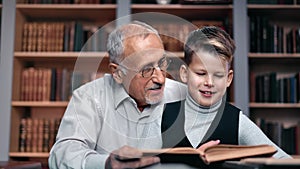 Happy 70s elderly grandfather and little schooler grandson reading book at library