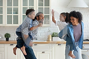 Happy active African parents piggybacking sibling children, running in kitchen