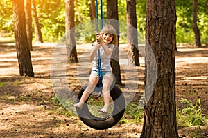 Happy active child girl playing on swing wheel in forest on sunny summer day. Preschool child having fun and swinging on a tire