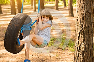 Happy active child girl playing on swing wheel in forest on sunny summer day. Preschool child having fun and swinging on a tire