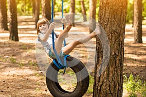 Happy active child girl playing on swing wheel in forest on sunny summer day. Preschool child having fun and swinging on a tire