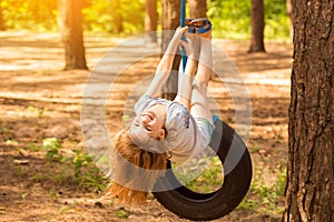 Happy active child girl playing on swing wheel in forest on sunny summer day. Preschool child having fun and swinging on a tire