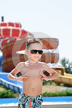 Happy active little handsome boy in sunglasses and shorts standing in summer aqua water amusement park sunbathing