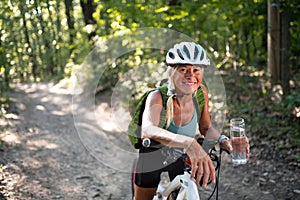 Happy active senior woman biker holding water bottle when sitting on bike outdoors in forest.