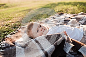 Happy adorable baby boy is lying on blanket on the grass in the park on summer day. Child in trendy and cute clothes