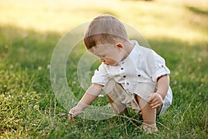 Happy adorable baby boy sitting on the grass in the park on summer day. Child in trendy and cute clothes