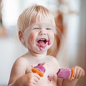 Happy adorable infant baby boy child smiling while eating two frozen fruit popsicle ice creams in simmer.