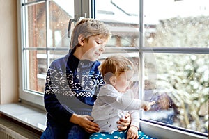 Happy adorable kid boy and cute baby girl sitting near window and looking outside on snow on Christmas day or morning