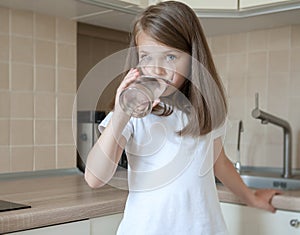 Happy adorable little girl drinking water in the kitchen at home. Caucasian kid  with long brown hair holding transparent glass
