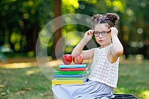 Happy adorable little kid girl reading book and holding different colorful books, apples and glasses on first day to