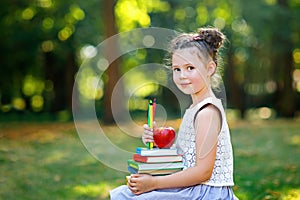 Happy adorable little kid girl reading book and holding different colorful books, apples and pencils on first day to