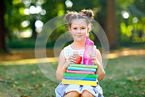 Happy adorable little kid girl reading book and holding different colorful books, apples and water bottle on first day