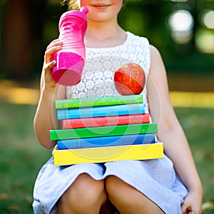 Happy adorable little kid girl reading book and holding different colorful books, apples and water bottle on first day