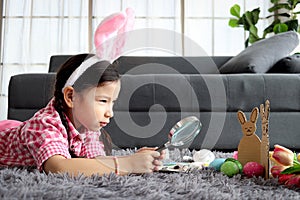 Happy adorable pink Easter bunny girl kid with rabbit ears headband holding magnifying glass for finding colorful painted Easter